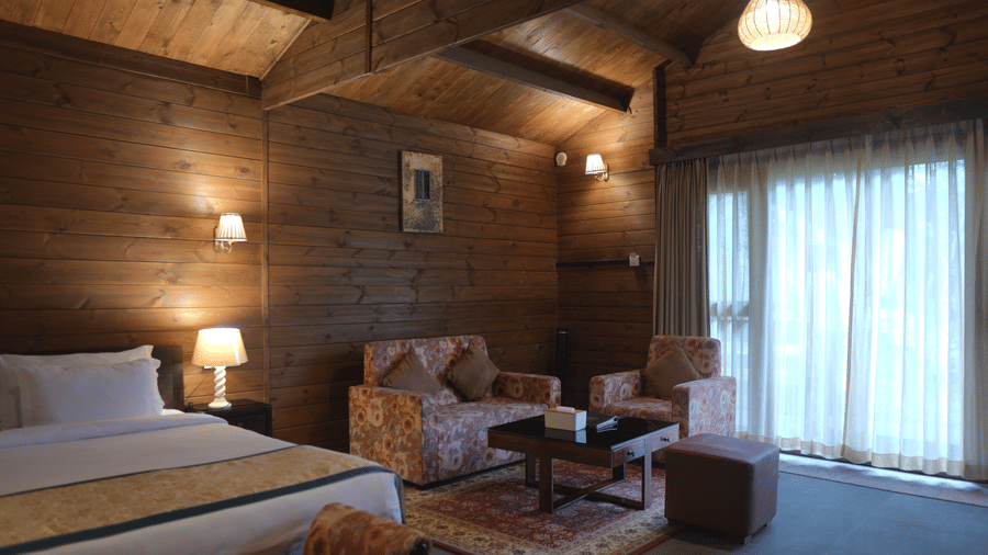 A bedroom at The Heritage Club Tripura Castle Hotel and Spa, Shillong featuring a double bed, a sofa set, a coffee table, wood panelled walls, and curtained windows.  