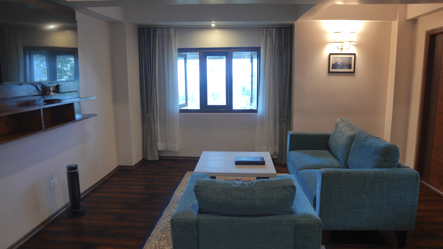 A living room at The Heritage Club Tripura Castle Hotel and Spa, Shillong featuring a sofa, an armchair, a coffee table, a window with curtains, and wall lights.