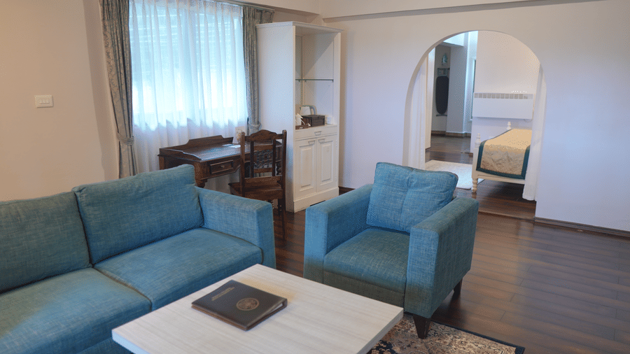 A living room at The Heritage Club Tripura Castle Hotel and Spa, Shillong featuring a sofa, an armchair, a coffee table, a study desk, and an arched doorway leading to a bedroom.