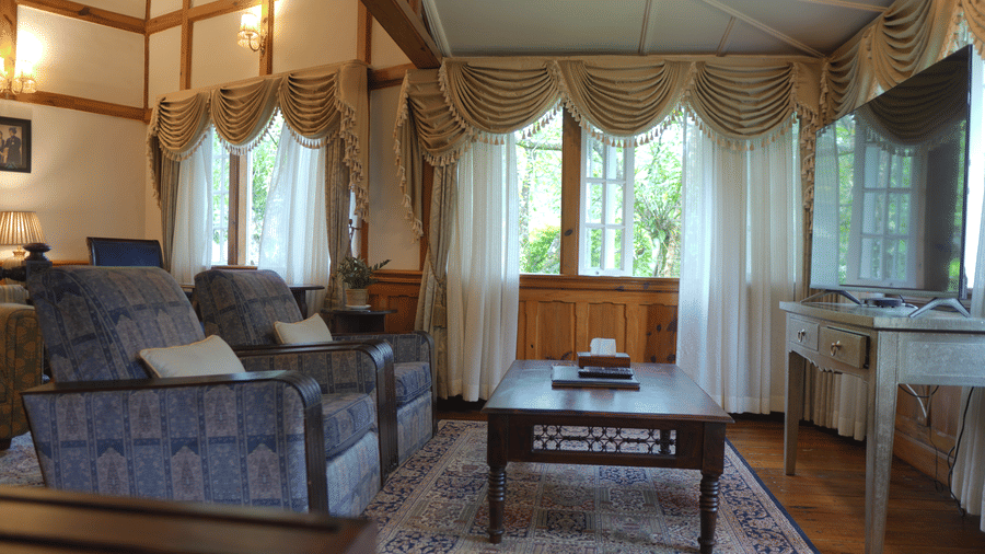 A living room at The Heritage Club Tripura Castle Hotel and Spa, Shillong featuring 2 armchairs, a coffee table, a television, a console table, and draped windows.  