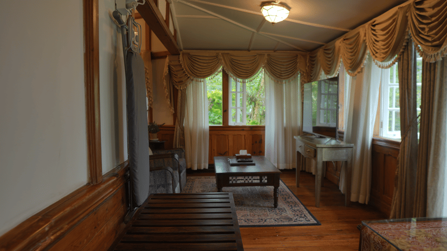 A living room at The Heritage Club Tripura Castle Hotel and Spa, Shillong featuring a seating area with armchairs, a coffee table, an ironing board, and draped windows.  