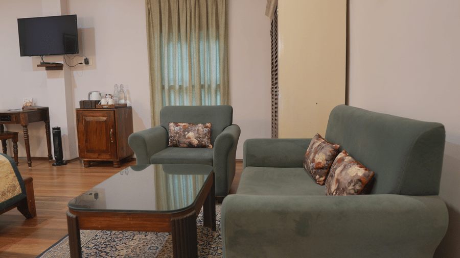 A living room at The Heritage Club Tripura Castle Hotel and Spa, Shillong featuring a sofa, an armchair, a coffee table, and a curtained window.  