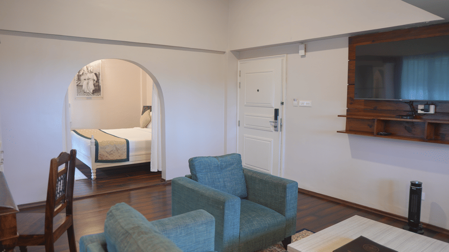 A living room at The Heritage Club Tripura Castle Hotel and Spa, Shillong featuring two armchairs, a coffee table, a wall-mounted television, and an arched entryway leading to the bedroom.  
