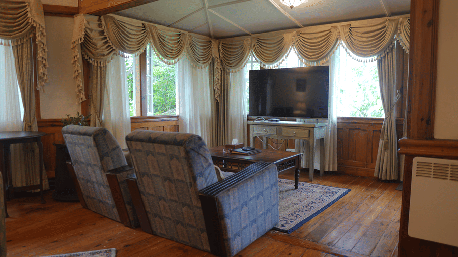 A living room at The Heritage Club Tripura Castle Hotel and Spa, Shillong featuring two plush armchairs, a coffee table, a television, and draped window valances.  
