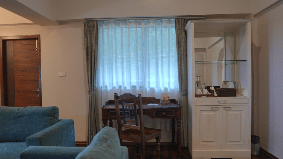 A room at The Heritage Club Tripura Castle Hotel and Spa, Shillong featuring a sofa, a wooden study desk, a chair, and a display cabinet near a curtained window.