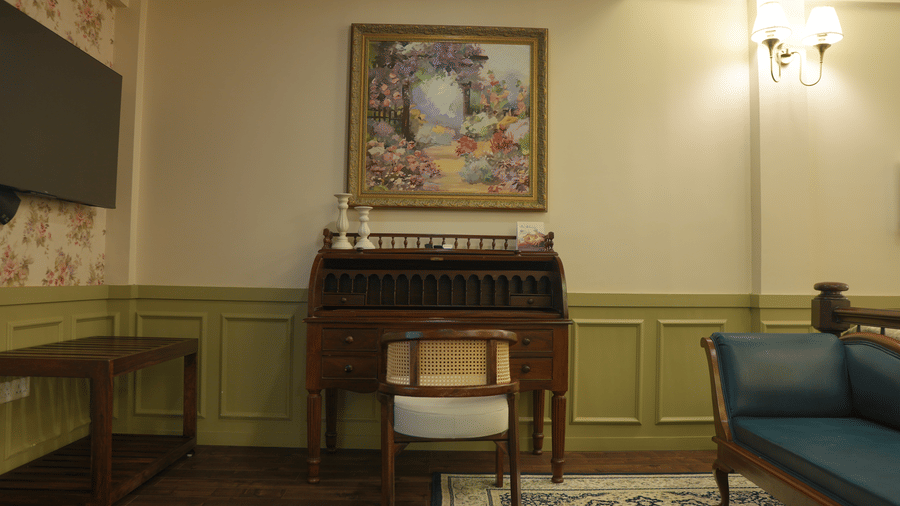A room at The Heritage Club Tripura Castle Hotel and Spa, Shillong featuring a wooden roll-top writing desk, a chair, wall art, and a blue settee sofa.  