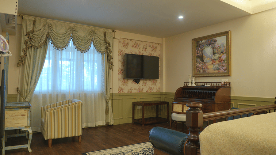 A room at The Heritage Club Tripura Castle Hotel and Spa, Shillong featuring an armchair, a writing bureau, a television, and draped window curtains.  