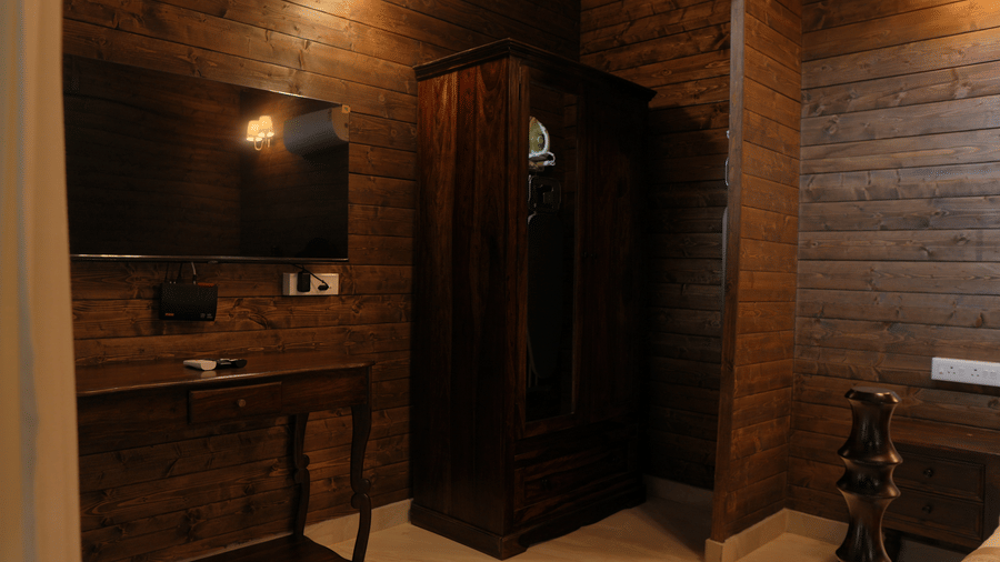 A room section at The Heritage Club Tripura Castle Hotel and Spa, Shillong featuring a wooden wardrobe, a console table with a wall-mounted television, and wood-panelled walls.  