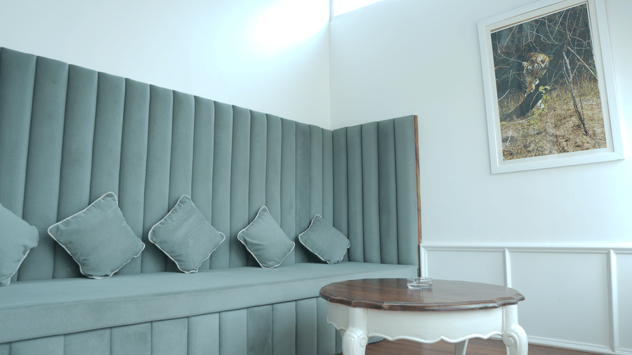 A seating area at The Heritage Club Tripura Castle Hotel and Spa, Shillong featuring a padded bench sofa, cushions, a round coffee table, and framed wall art.  