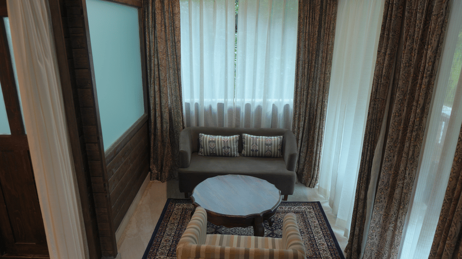 A sitting area at The Heritage Club Tripura Castle Hotel and Spa, Shillong featuring a sofa, an armchair, a coffee table, a rug, and curtains.  