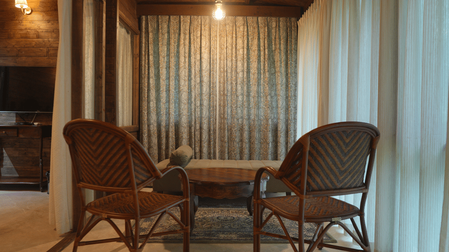 A sitting area at The Heritage Club Tripura Castle Hotel and Spa, Shillong featuring 2 wicker chairs, a daybed, a coffee table, and full-length window curtains.  