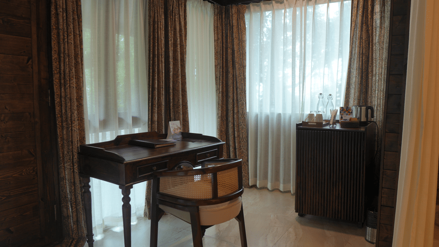A study area at The Heritage Club Tripura Castle Hotel and Spa, Shillong featuring a wooden writing desk, a chair, curtained windows, and a tea station cabinet.  