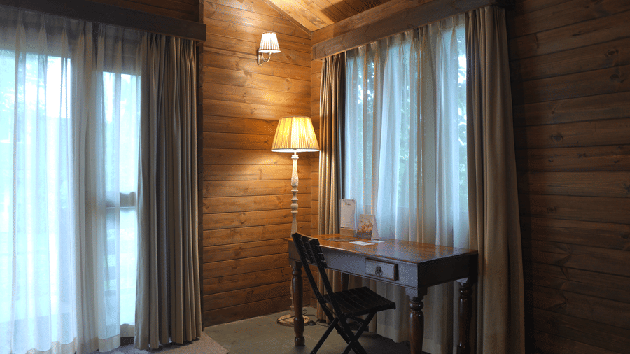 A study area at The Heritage Club Tripura Castle Hotel and Spa, Shillong featuring a wooden writing desk, a chair, a floor lamp, and curtained windows.  
