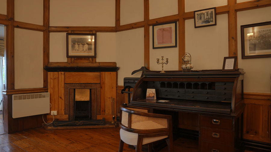 A room at The Heritage Club Tripura Castle Hotel and Spa, Shillong featuring a wooden roll-top desk, a chair, a fireplace, and framed picture frames.  