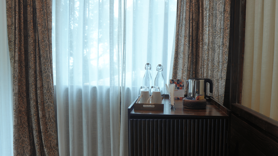 A tea station at The Heritage Club Tripura Castle Hotel and Spa, Shillong featuring an electric kettle, water bottles, cups, and a wooden cabinet set by a curtained window.  