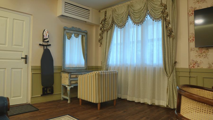 A room at The Heritage Club Tripura Castle Hotel and Spa, Shillong featuring a vanity desk with a mirror, an armchair, an ironing board, and draped curtains.  