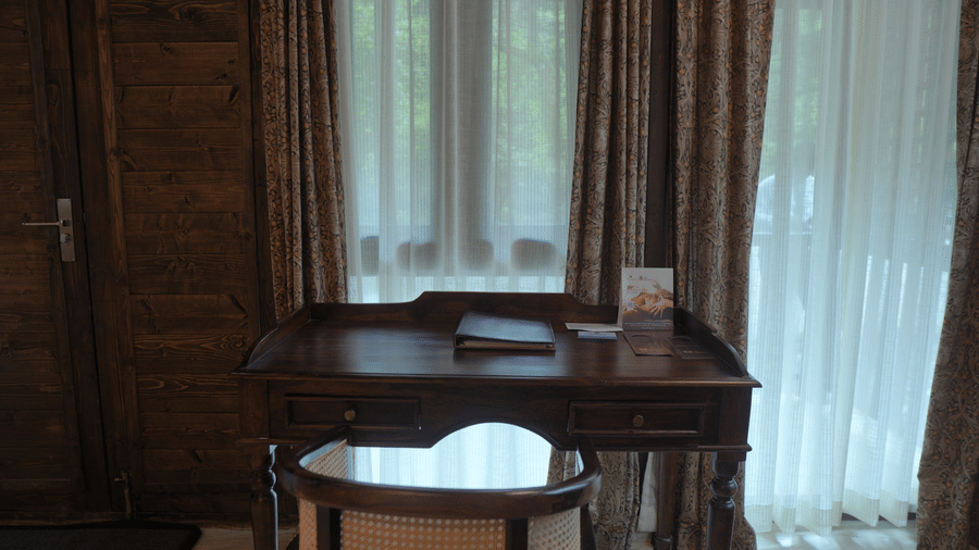 A wooden study desk with a chair in front of draped windows at The Heritage Club Tripura Castle Hotel and Spa, Shillong.