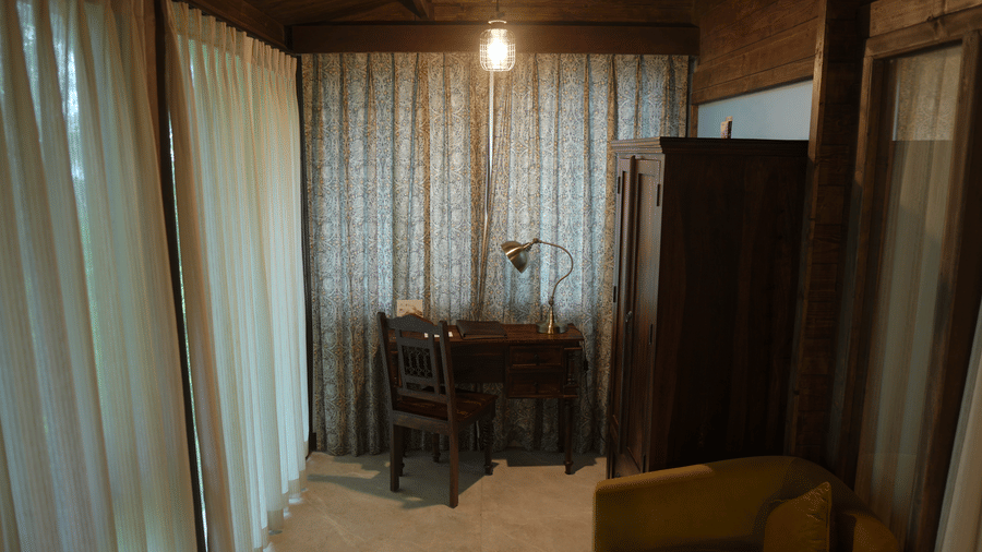 A work corner at The Heritage Club Tripura Castle Hotel and Spa, Shillong featuring a wooden writing desk, a chair, a desk lamp, a wardrobe, and curtained windows.  
