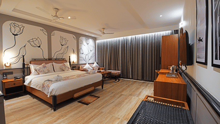 Corner view of the Suite at Hotel Sonar Bangla, Kolaghat, featuring wooden flooring, a double bed facing a wall-mounted TV, and a seating area beside closed curtains.