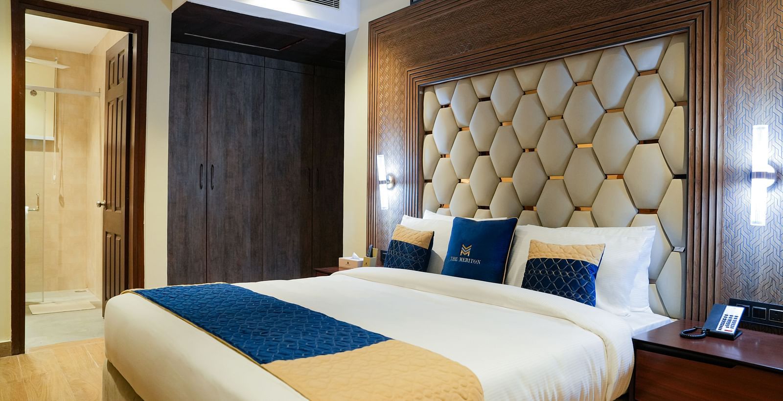 A well-appointed double bedroom at The Meriton, featuring a large bed with a padded headboard, crisp white and blue bedding, and a bedside table with a lamp at the Empress Suite.