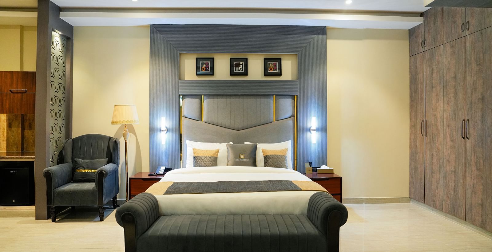 A spacious and elegantly designed bedroom of a Premium Room at The Meriton, featuring a large bed with a padded headboard, a chaise lounge at the foot of the bed, and warm lighting.