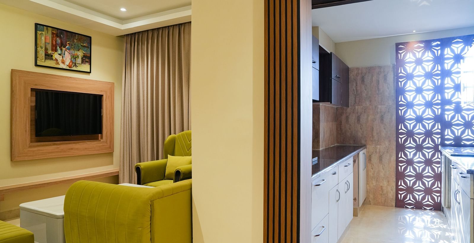 Corner view of a bright and inviting living area of the Junior Suite in The Meriton, featuring lime green sofas, a wall-mounted television, and a glimpse of an adjoining room with patterned curtains.