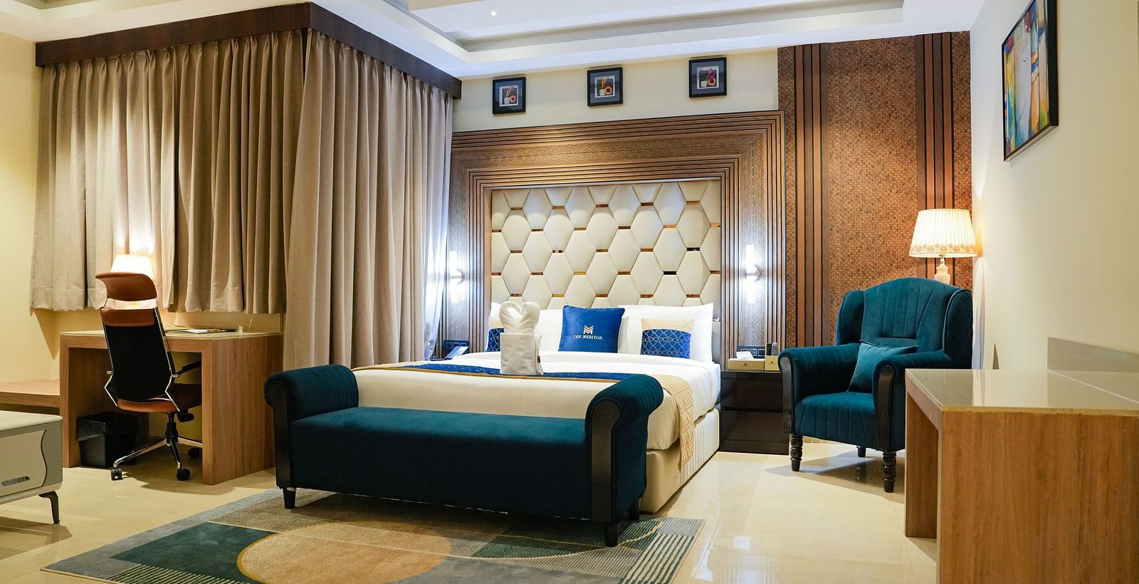 A luxurious bedroom in the Presidential Suite at The Meriton, featuring a large king size bed with a padded headboard, a stylish blue chaise lounge, and an armchair with a side table and lamp.