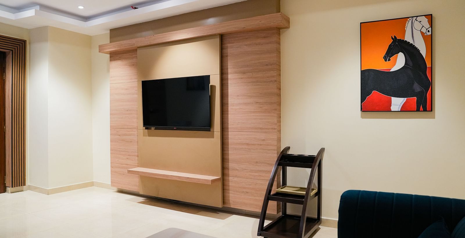A modern living area in the Presidential Suite at The Meriton, featuring a wall-mounted television integrated into light wooden panelling, a dark sofa, and a vibrant abstract painting on the wall.