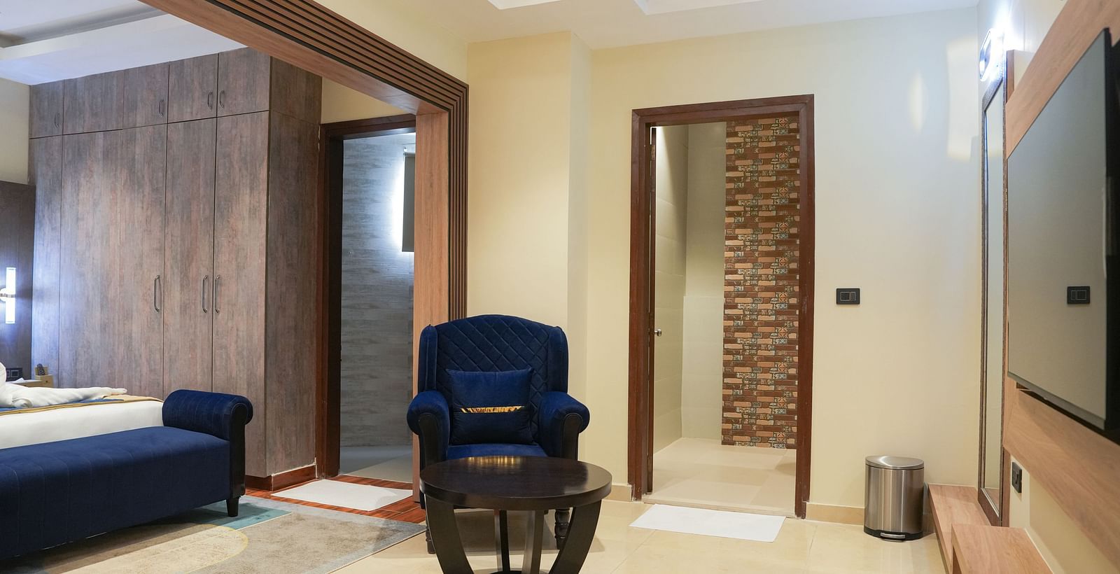 A partial view of the Standard Room at The Meriton, highlighting a blue chaise lounge, a wooden bedside table, and the entrance to a bathroom with a glass shower door visible at the far end.