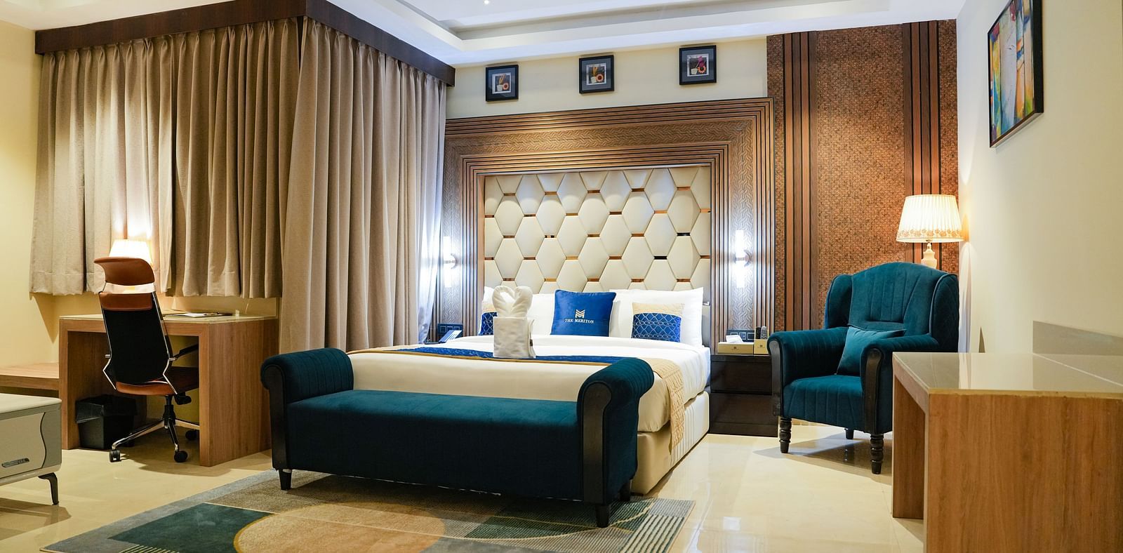 A luxurious bedroom in the Presidential Suite at The Meriton, featuring a large king size bed with a padded headboard, a stylish blue chaise lounge, and an armchair with a side table and lamp.