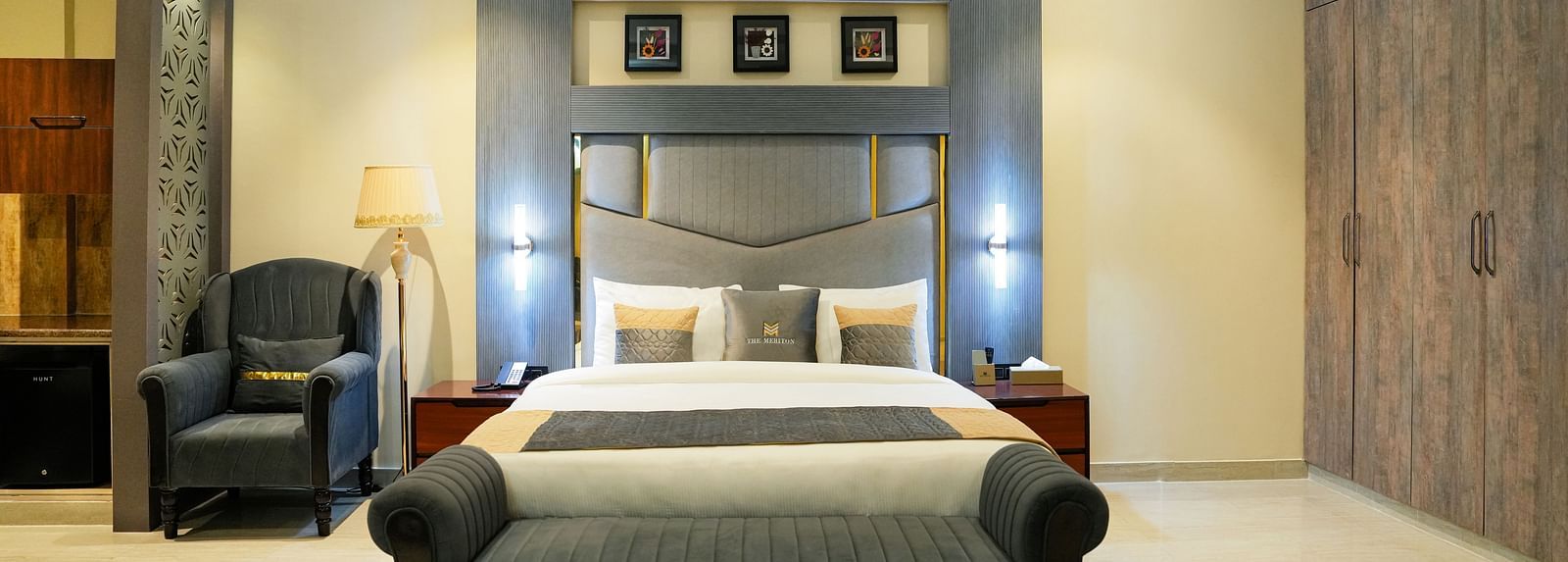 A spacious and elegantly designed bedroom of a Premium Room at The Meriton, featuring a large bed with a padded headboard, a chaise lounge at the foot of the bed, and warm lighting.