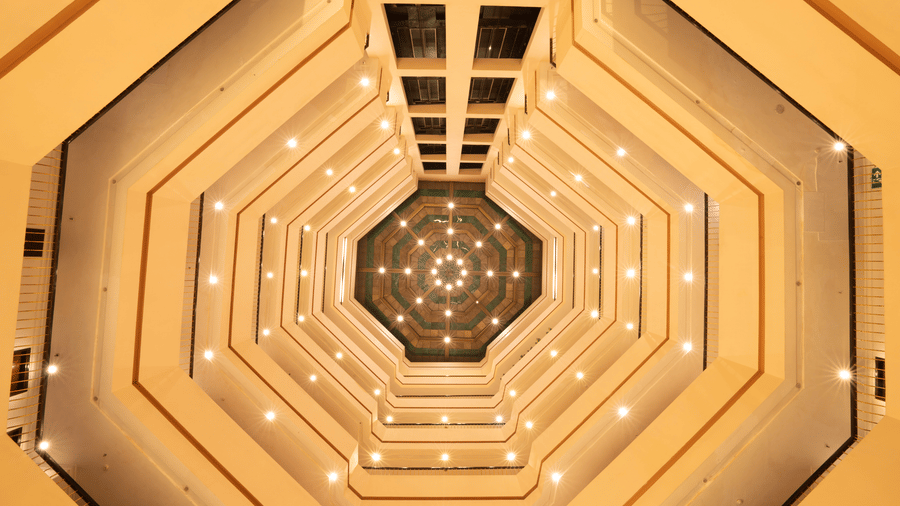 An upward perspective of an octagonal indoor atrium with multiple floor levels and overhead lighting | HHI Varanasi