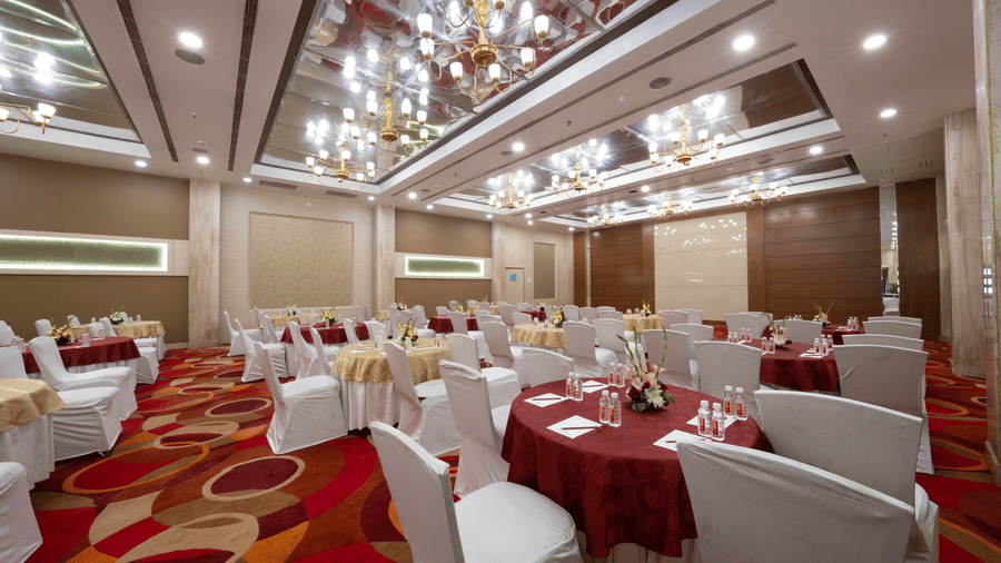 A large hall with round tables covered in red and white cloths, white chairs, and patterned carpeting | HHI Varanasi