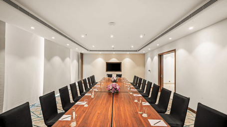 Grand Club Lounge I in Vijayawada with a boardroom-style meeting setup, wooden conference table, comfortable seating, and a display screen | Grand Vijayawada by GRT Hotels
