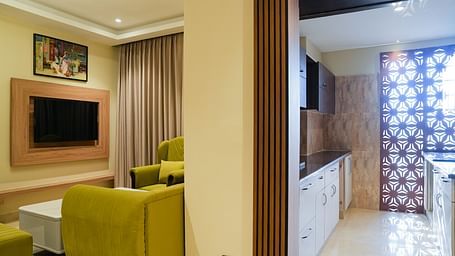 Corner view of a bright and inviting living area of the Junior Suite in The Meriton, featuring lime green sofas, a wall-mounted television, and a glimpse of an adjoining room with patterned curtains.