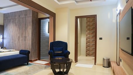 A partial view of the Standard Room at The Meriton, highlighting a blue chaise lounge, a wooden bedside table, and the entrance to a bathroom with a glass shower door visible at the far end.