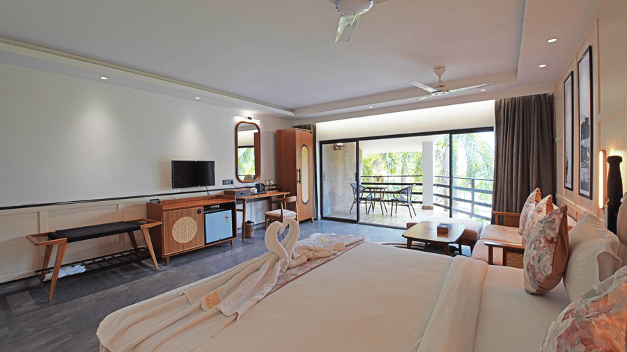 Side view of the Premium Room at Hotel Sonar Bangla, Kolaghat, featuring its double bed facing a TV, curtains drawn to reveal the balcony with outdoor seating.