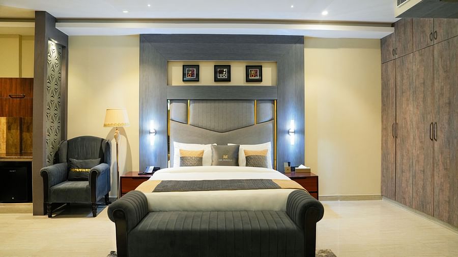 A spacious and elegantly designed bedroom of a Premium Room at The Meriton, featuring a large bed with a padded headboard, a chaise lounge at the foot of the bed, and warm lighting.