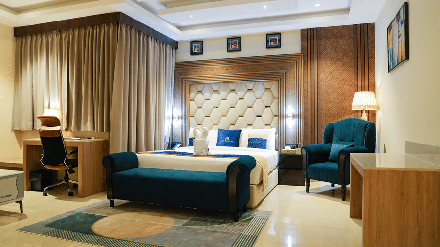 A luxurious bedroom in the Presidential Suite at The Meriton, featuring a large king size bed with a padded headboard, a stylish blue chaise lounge, and an armchair with a side table and lamp.