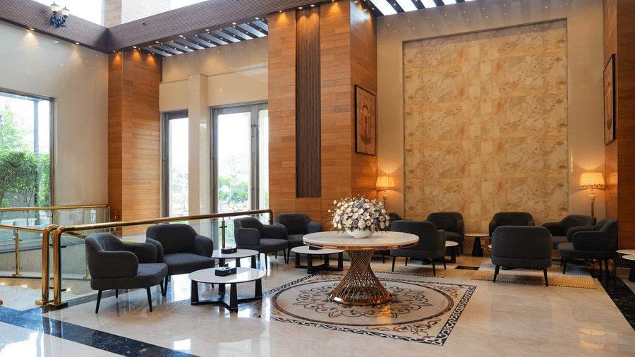 A wide view of our hotel near Nehru Place's sophisticated common area, featuring dark upholstered seating, round tables, and decorative wall panels | Location of The Meriton Hotel, New Delhi
