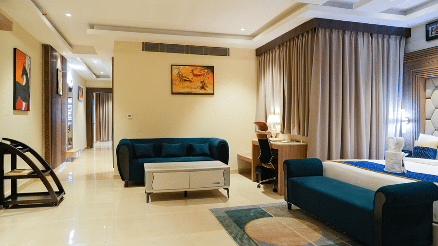 A comfortable and well-appointed living room area in a Presidential Suite in Delhi at The Meriton, complete with a teal sofa, a coffee table, and an armchair, with artistic prints on the walls.