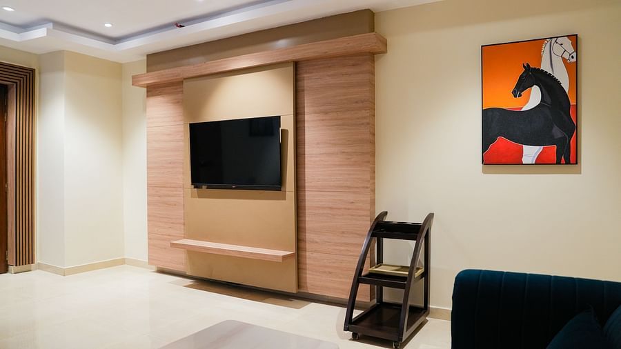 A modern living area in the Presidential Suite at The Meriton, featuring a wall-mounted television integrated into light wooden panelling, a dark sofa, and a vibrant abstract painting on the wall.