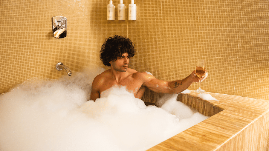 A shirtless man with curly hair relaxing in a luxury sunken bubble bath while holding a drink glass in a warm-toned resort bathroom.