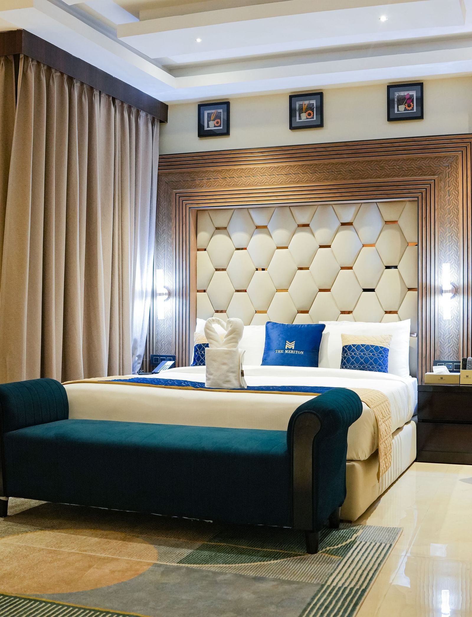 A luxurious bedroom in the Presidential Suite at The Meriton, featuring a large king size bed with a padded headboard, a stylish blue chaise lounge, and an armchair with a side table and lamp.