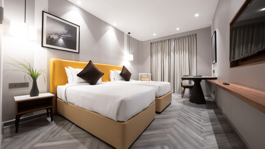 Twin room at The HHI Varanasi featuring 2 beds with yellow headboard and white bedding with dark accent pillows and bedside table alongside a work desk and wall-mounted TV with sheer curtains.
