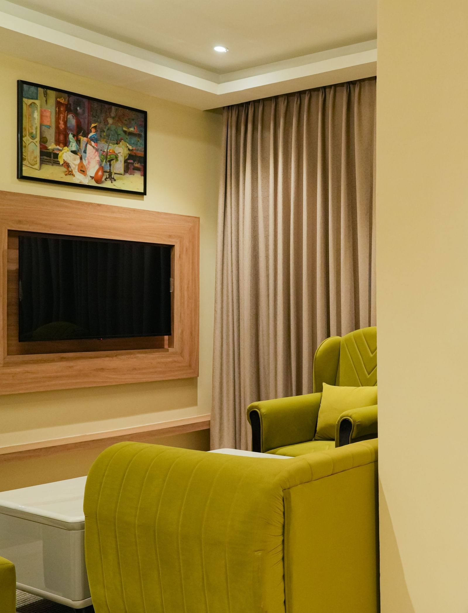 Corner view of a bright and inviting living area of the Junior Suite in The Meriton, featuring lime green sofas, a wall-mounted television, and a glimpse of an adjoining room with patterned curtains.