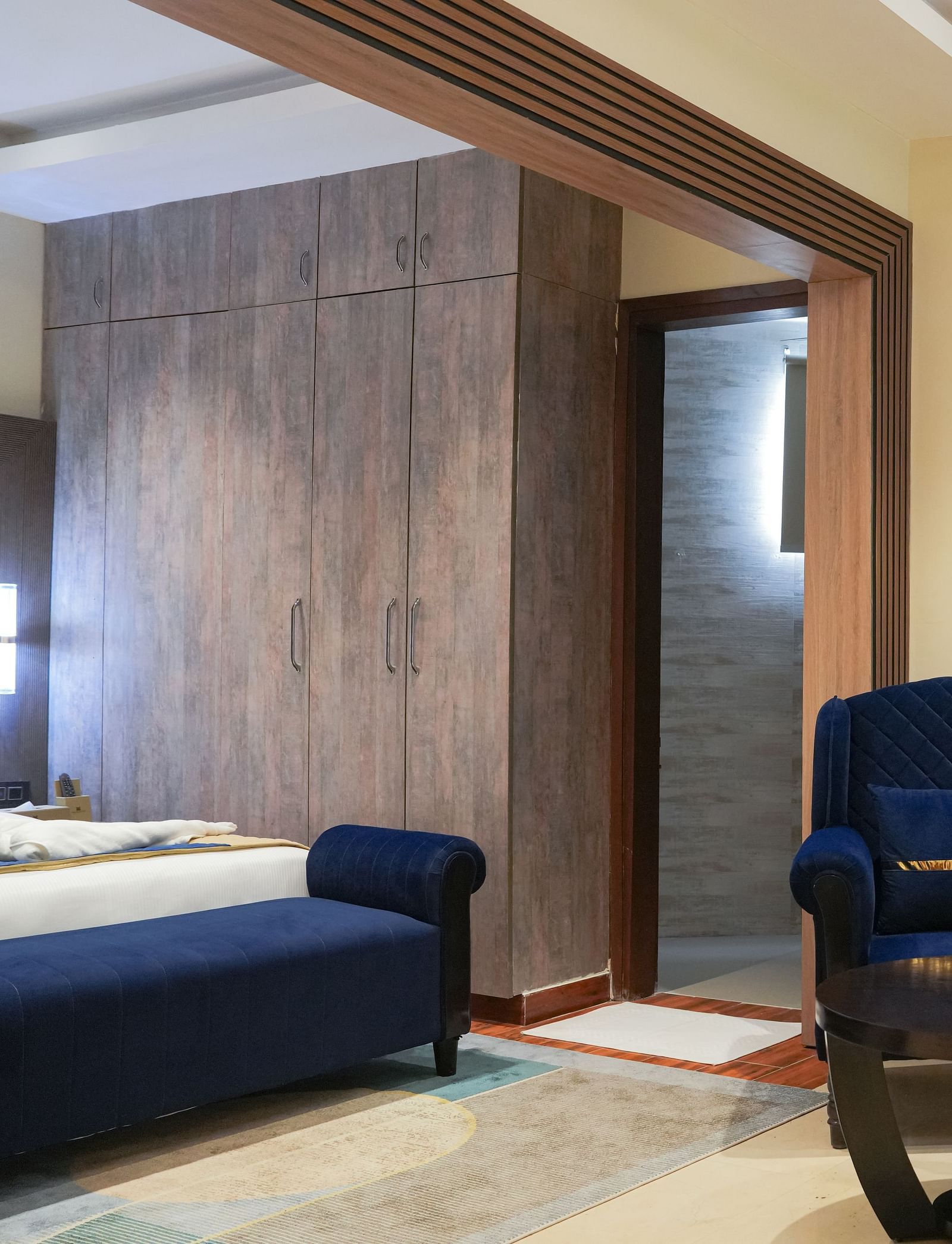 A partial view of the Standard Room at The Meriton, highlighting a blue chaise lounge, a wooden bedside table, and the entrance to a bathroom with a glass shower door visible at the far end.