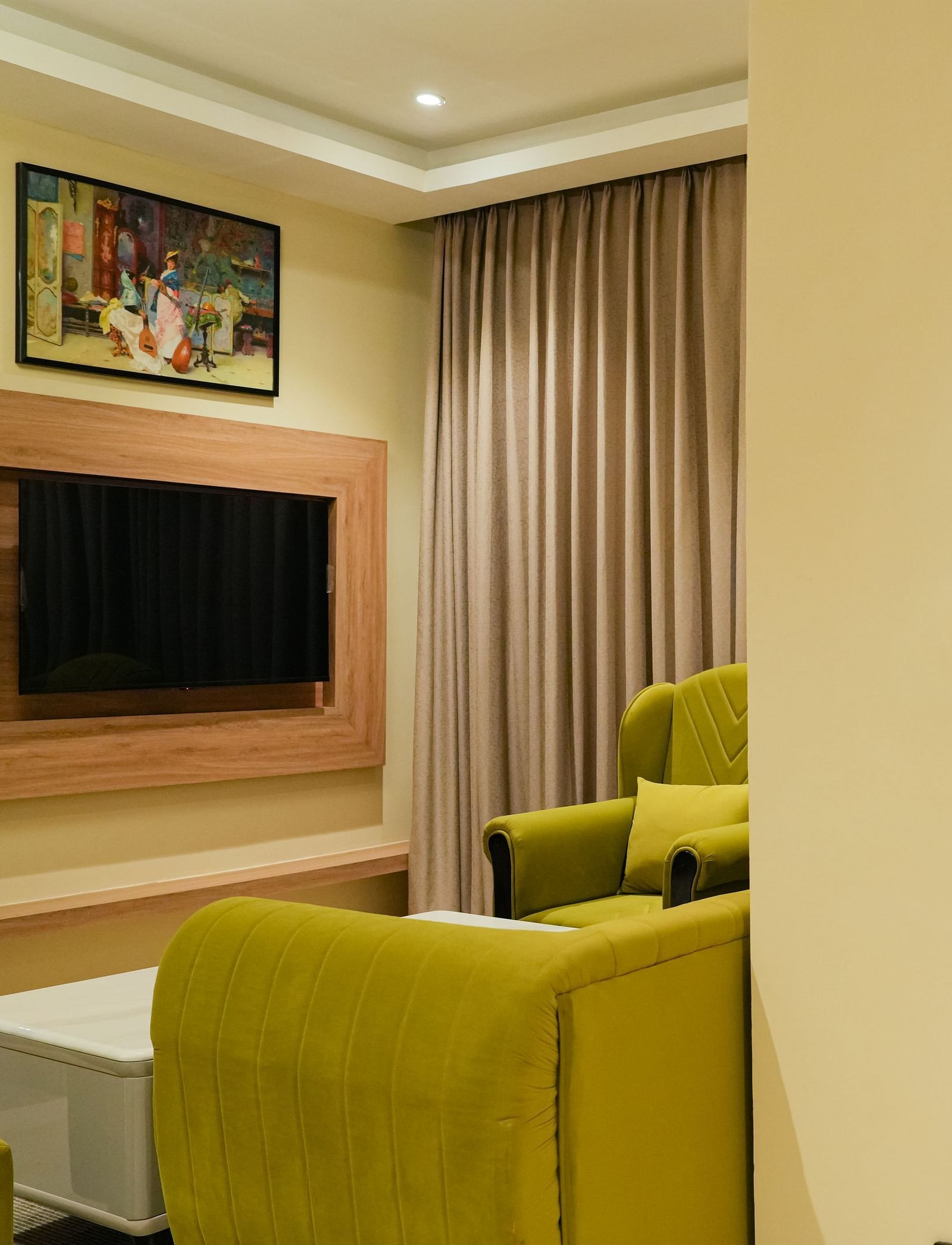 Corner view of a bright and inviting living area of the Junior Suite in The Meriton, featuring lime green sofas, a wall-mounted television, and a glimpse of an adjoining room with patterned curtains.