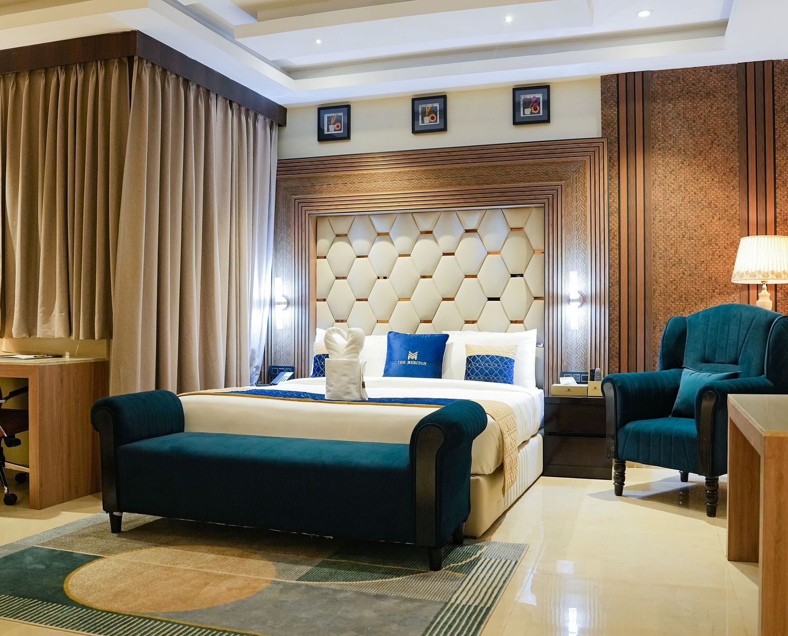 A luxurious bedroom in the Presidential Suite at The Meriton, featuring a large king size bed with a padded headboard, a stylish blue chaise lounge, and an armchair with a side table and lamp.
