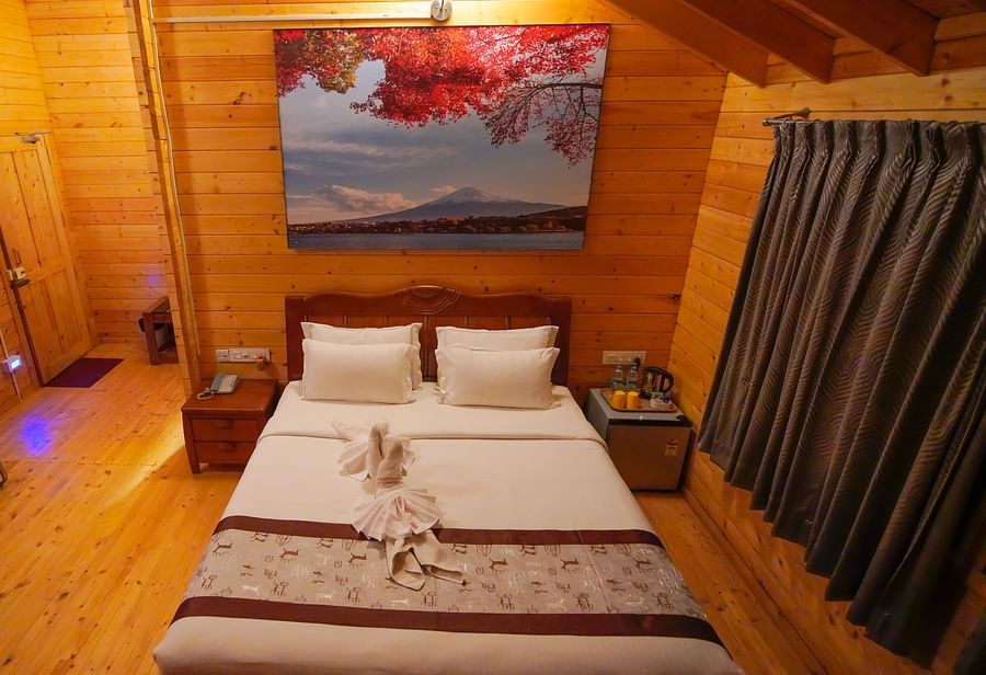 Wooden House of GK Hill View Resort furnished with a double bed, seating bench, bedside tables, overhead lighting, wooden architecture and curtained windows.